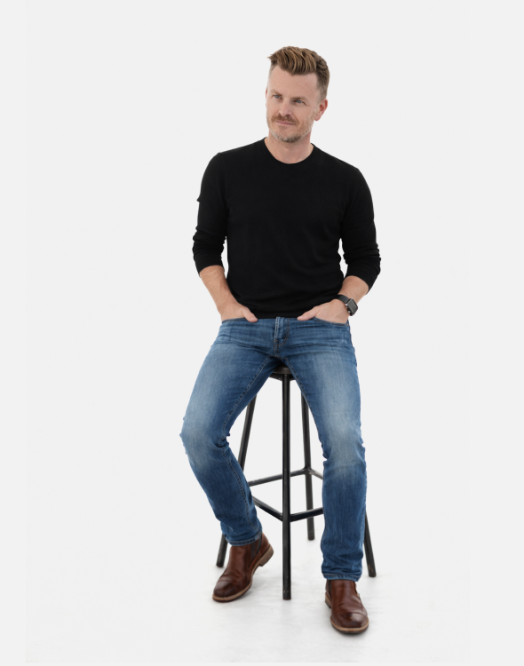 photo of Danny sitting on stool. Danny wearing jeans and a black v-neck sweater staring off to left about 30 degrees