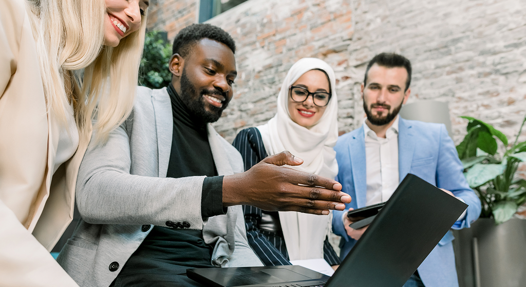 Team of multiethnical diverse business people, two men and two women, standing in modern office, discussing and working together, using laptop and tablet. Business and digital.